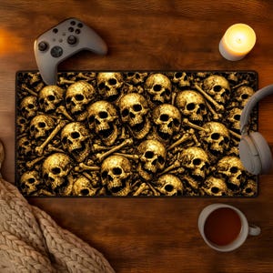 May include: A rectangular mouse mat with a repeating pattern of gold skulls and bones. A grey game controller, headphones, and a lit candle are on the wooden surface. A cup of tea and a knitted blanket are also visible.