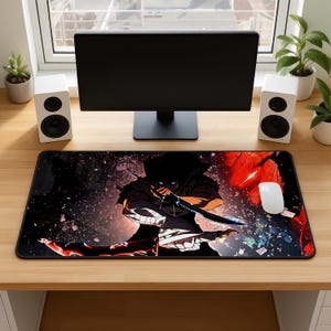 May include: A rectangular desk mat featuring a dark, stylized illustration of a hooded figure wielding a glowing blue blade. The artwork includes vibrant red accents, scattered debris, and a black background. A white computer mouse rests on the mat.