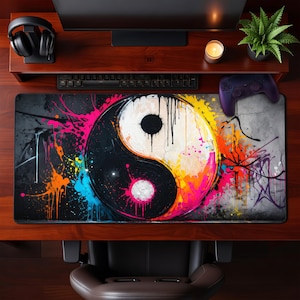 Puede incluir: Una alfombrilla de escritorio rectangular grande con un diseño de Yin y Yang. El diseño está dividido en mitades blancas y negras, con salpicaduras de pintura coloridas en rosa, naranja, amarillo y azul. La alfombrilla está sobre un escritorio de madera con teclado, auriculares y un mando.