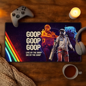 以下が含まれることがあります： 「GOOP GOOP GOOP」と「LIVE BY THE GOOP DIE BY THE GOOP」のテキストが入ったゲーミングマウスパッドは、グレーのコントローラー、ヘッドフォン、ろうそく、紅茶のカップに囲まれています。マウスパッドは、2つのフィギュアを使ったカラフルなデザインです。