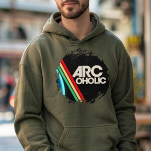 Puede incluir: Una sudadera con capucha de color verde oliva que presenta un gr&aacute;fico circular en el pecho. El dise&ntilde;o incluye un c&iacute;rculo negro desgastado con el texto blanco ARC OHOLIC y tres rayas diagonales en azul, amarillo y rojo. Tiene un bolsillo frontal.