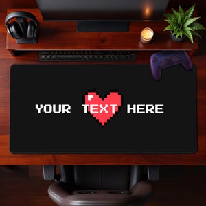 Peut inclure: Un tapis de bureau rectangulaire noir orné d'un cœur rouge pixélisé centré entre le texte blanc pixélisé qui indique YOUR TEXT HERE. Le tapis présente une surface lisse et des coins arrondis, conçu pour être placé sous un clavier et une souris.