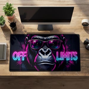 May include: A black rectangular desk mat with a gorilla wearing pink sunglasses. The words "OFF LIMITS" are written in neon blue and pink. The desk mat is on a wooden desk with a computer monitor and plants.