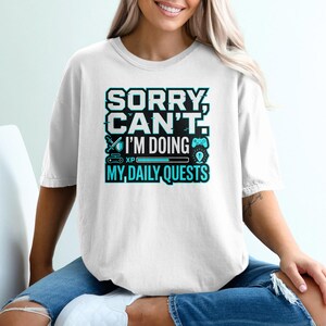 May include: White t-shirt with the text "SORRY, CAN'T. I'M DOING MY DAILY QUESTS" in a teal and black design. The design includes a sword, game controller, and a progress bar. The shirt is worn by a person.