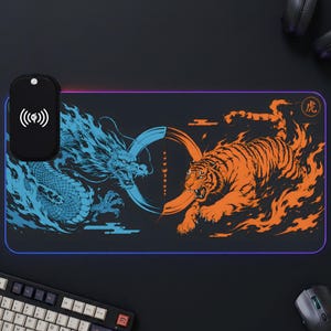 Könnte beinhalten: Eine rechteckige schwarze Schreibtischunterlage mit einem blauen Drachen auf der linken und einem orangefarbenen Tiger auf der rechten Seite, getrennt durch ein zentrales kreisförmiges Motiv. Ein schwarzes kabelloses Ladepad ist links angebracht.
