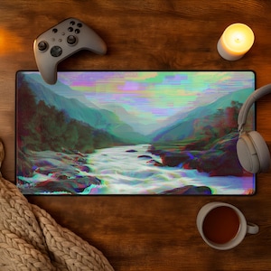 Peut inclure: Tapis de souris rectangulaire avec un paysage d'art num&eacute;rique d'une rivi&egrave;re et de montagnes, dans des tons de vert, de bleu et de rouge. Une manette de jeu grise et un casque sont sur la surface en bois, avec une bougie allum&eacute;e et une tasse de th&eacute;.