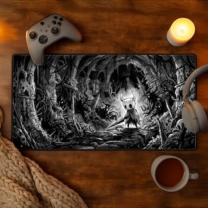 May include: A rectangular desk mat featuring a black and white illustration of a small, horned character holding a needle-like sword in a dark, cavernous environment with twisted roots and ruins, surrounded by a game controller and headphones.