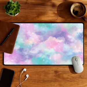 May include: A rectangular mouse pad with a colorful galaxy design in shades of pink, purple, and teal. The pad is surrounded by a black border. A white mouse, a smartphone, and a pair of headphones are on the wooden surface.