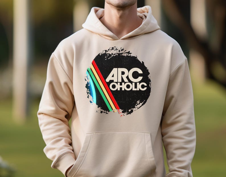Puede incluir: Una sudadera con capucha de color beige que presenta un dise&ntilde;o gr&aacute;fico central. El dise&ntilde;o consiste en un fondo circular negro con aspecto desgastado, el texto blanco ARC OHOLIC y tres rayas diagonales en azul, amarillo y verde.