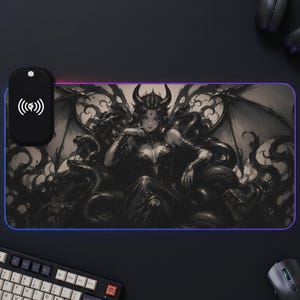 Puede incluir: Una alfombrilla de ratón rectangular para juegos con una ilustración oscura y detallada de una figura con cuernos rodeada de serpientes y alas. La alfombrilla incluye un módulo de carga inalámbrica negro con un icono de símbolo Qi blanco.