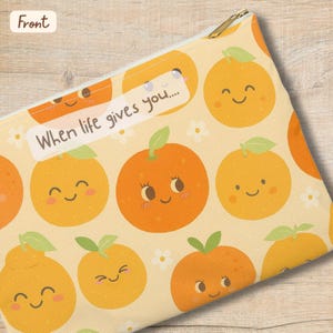 Puede incluir: Estuche rectangular con un patrón repetido de naranjas sonrientes sobre un fondo amarillo claro. El texto "When life gives you..." está impreso en un rectángulo blanco. El estuche tiene una cremallera dorada.