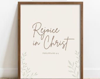 Könnte beinhalten: Ein gerahmter Kunstdruck mit dem Text "Rejoice in Christ" in brauner Schreibschrift und "PHILIPPIANS 4:4" in einer kleineren Schriftart darunter. Das Design zeigt hellgraue Blattillustrationen in den unteren Ecken auf einem cremefarbenen Hintergrund.