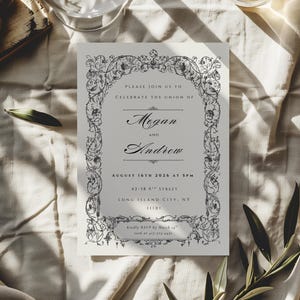 May include: A wedding invitation with an ornate floral border. The invitation features the names "Megan and Andrew" and the date "August 16th, 2026". The invitation is on a cream-colored paper, with the text in a dark gray color.