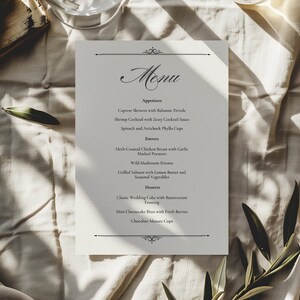 Könnte beinhalten: Eine gedruckte Menükarte mit dem Wort „Menü“ in eleganter Schrift oben. Die Speisekarte listet Vorspeisen, Hauptgerichte und Desserts auf. Die Karte befindet sich auf einer strukturierten, cremefarbenen Oberfläche mit dekorativen Elementen oben und unten.