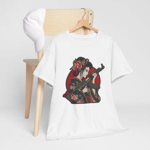 Könnte beinhalten: Weißes T-Shirt mit einer Grafik einer Geisha, die ein Gewehr vor rotem Hintergrund hält. Die Geisha trägt florale Haaraccessoires und einen traditionellen Kimono. Eine weiße Baseballkappe liegt auf einem Holzstuhl.