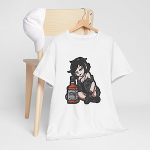 May include: White t-shirt with a cartoon illustration of a person with black hair and clothing, sitting beside a Jack Daniel's whiskey bottle. The shirt is on a wooden hanger.