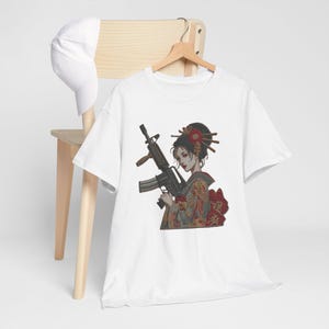 May include: White t-shirt with a graphic of a woman in Japanese traditional dress holding a rifle. The illustration has red and gold details in the woman's clothing and hair. The shirt is displayed on a wooden hanger.