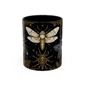 May include: Black ceramic mug featuring detailed illustrations of insects in gold and white. The design includes a large moth, a beetle, and other insect motifs against a dark background. The mug has a black interior and rim.