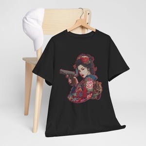 May include: Black t-shirt featuring a graphic of a woman in a kimono holding a handgun. The illustration has a Japanese aesthetic with floral and traditional design elements. A white baseball cap hangs on a wooden chair.