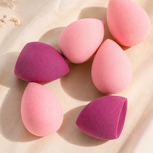 Può includere: Un assortimento di sei spugne per il trucco in tonalità rosa e viola. Le spugne sono a forma di goccia e angolate, progettate per applicare trucco liquido o in crema. Lo sfondo è un tessuto morbido e neutro.