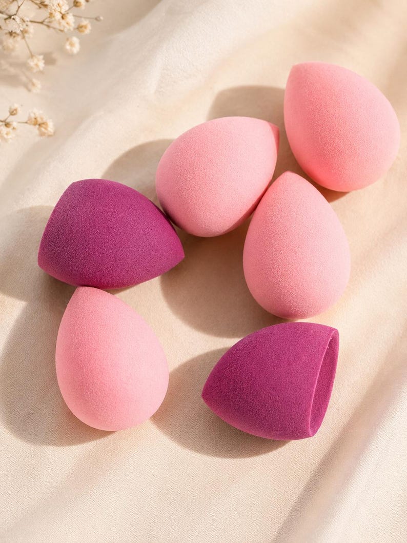 May include: A selection of six makeup sponges in pink and purple hues. The sponges are teardrop-shaped and angled, designed for applying liquid or cream makeup. The background is a soft, neutral fabric.