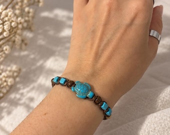 Puede incluir: Una pulsera de cordón marrón con un dije central de tortuga de color turquesa, acentuada con pequeñas cuentas cuadradas turquesas y cuentas redondas marrones alternadas, asegurada con un diseño de nudos.