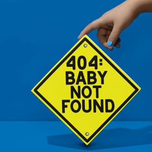 May include: A yellow diamond sign with black border and the text "404: BABY NOT FOUND". The sign is held by a hand with blue nail polish against a blue background.