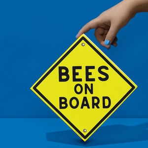 May include: A yellow diamond-shaped sign with black text that reads "BEES ON BOARD." The sign has a black border and is held by a hand with blue nail polish against a blue background. The sign has two small silver rivets.