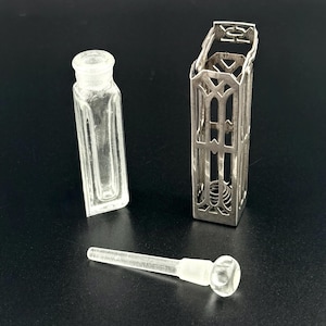 May include: A clear glass perfume bottle with a rectangular shape and a separate glass stopper. A decorative silver metal case with an openwork design surrounds the bottle. The case has a hinged top and a detailed pattern.