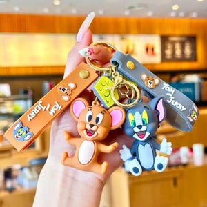 May include: Keychains featuring cartoon characters Tom and Jerry. One keychain has a brown Jerry figure, and the other has a gray Tom figure. Both have matching straps with the words "Tom" and "Jerry" printed on them. The keychains are attached to a gold-colored ring.