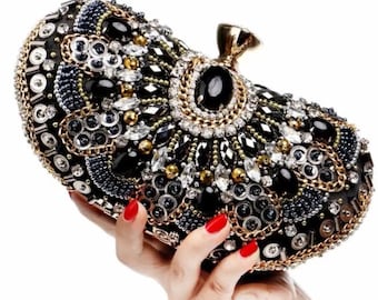 May include: An ornate black clutch with gold, silver, and black embellishments. The evening bag features a large black stone at the center, surrounded by a gold chain and smaller crystals. The clutch is held by a hand with red nail polish.