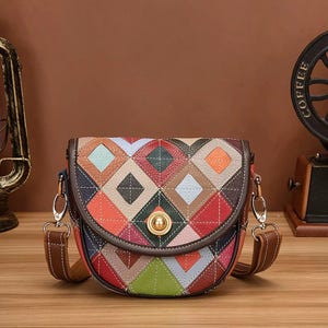 May include: A colorful, patchwork crossbody bag with a geometric diamond pattern. The bag features a brown leather strap and a gold-colored clasp. The bag is made of various colors including red, orange, green, and blue.