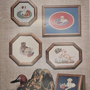 May include: A collection of framed cross-stitch art featuring sporting puppies. The artwork includes various dog breeds in different scenes, such as playing with toys or near hunting gear. The title "Pick of the Litter" is displayed at the top.