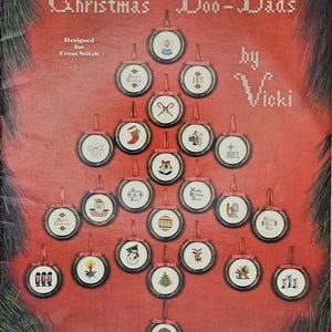 May include: A vintage Christmas cross-stitch pattern book titled "Christmas Doo-Dads" by Vicki. The cover features a Christmas tree design made of round ornaments with various holiday-themed cross-stitch designs. The background is red.