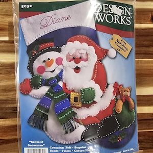 May include: A Design Works felt stocking craft kit featuring a Santa and snowman design. The package includes felt, sequins, beads, trims, cotton floss, a needle, and instructions to create a 16-inch stocking. The front displays the text Santa & Snowman.