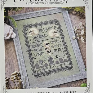 May include: A framed cross-stitch sampler with a Halloween theme. The design features skulls, tombstones, and the text "HALLOWED BE THY BONES." The sampler includes the alphabet, numbers, and the text "JN 25." The frame is gray.