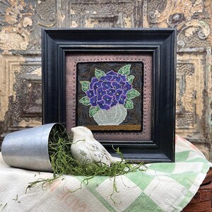 May include: A framed floral artwork features a bouquet of purple flowers in a white vase. The artwork is in a black frame, with a decorative bird and a metal container with greenery in the foreground. The background has a rustic, vintage aesthetic.