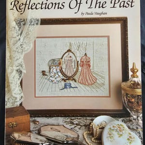 May include: A cross-stitch pattern booklet titled "Reflections Of The Past" by Paula Vaughan. The cover features a framed cross-stitch design of two women in a room with a mirror and a vanity table. The booklet is part of a series called "Leisure Arts Book Five".