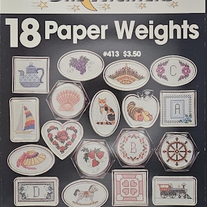May include: A vintage craft pattern book titled "One Nighters" featuring 18 paper weight designs. The cover is black with a crescent moon and star graphic. The designs include a teapot, grapes, a cat, and a sailboat, all in cross-stitch.