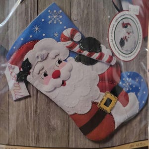May include: A Bucilla 150th Anniversary Special Edition felt stocking kit featuring a Santa Claus design with a red hat, white beard, and candy cane. The kit includes stamped felts, cotton floss, sequins, beads, and needles for an 18-inch project.