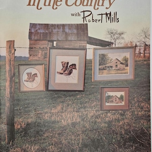 May include: A book cover titled "In the Country with Robert Mills" featuring framed artwork of country scenes and boots hanging on a barbed wire fence. The background shows a weathered barn and a grassy field.