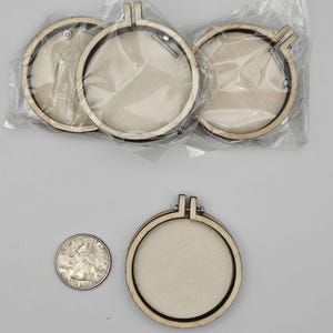 May include: A collection of wooden embroidery hoops, some packaged in clear plastic. The hoops are round with a light wood finish and a metal clasp. A US quarter is included for size comparison. The hoops are designed for embroidery or cross-stitch projects.