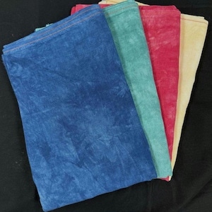May include: Four folded cloths in shades of blue, teal, red, and cream. The cloths have a textured appearance, with visible stitching along the edges. The blue cloth is in the foreground, with the other colors fanning out behind it. The cloths are arranged on a black surface.