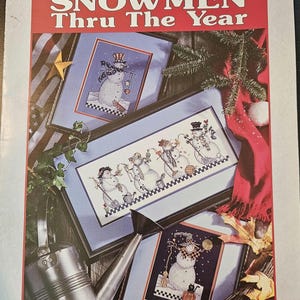 May include: A book cover with the title "Snowmen Thru The Year" by "goodnaturedgirls". The cover features three framed cross-stitch patterns of snowmen in different seasonal settings.