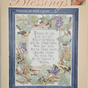 May include: A counted cross-stitch kit titled "Blessings" features a framed design with a religious verse, birds, and grape vines. The design is by Nancy Rossi and produced by Kooler Design Studio, Inc. The color palette includes shades of blue, green, purple, and yellow.