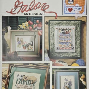 May include: A book cover for "CATS Galore" cross-stitch patterns. The cover features various framed cross-stitch designs of cats, including a design that says "I ❤️ CATS". The cover also includes the text "CHARTS FOR LESS" and "66 DESIGNS".