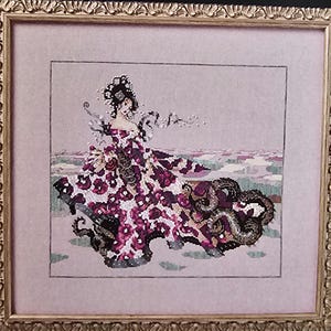 May include: Cross-stitch artwork of a woman in a flowing gown with a floral pattern in shades of purple and white. The artwork is framed in a gold-colored ornate frame. The background features a landscape in muted tones.