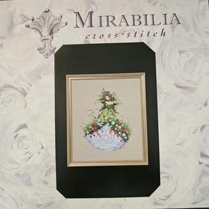 May include: A framed cross-stitch piece featuring a woman in a floral gown. The dress is adorned with red berries, white flowers, and green foliage. The background is a neutral beige, and the frame is gold. The words "Mirabilia cross-stitch" are at the top.