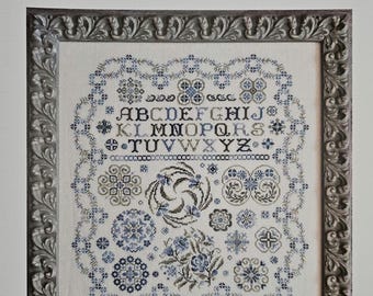 May include: Cross-stitch pattern titled "Serendipity" by Karen Kluba. The design features an alphabet, floral and geometric motifs in shades of blue and olive green. The pattern is framed in a decorative silver-colored frame.