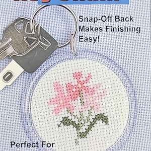 May include: Clear key chain with a cross-stitched pink flower design. The key chain has a silver ring and a key attached. Text on the packaging reads "Key Chain" and "Snap-Off Back Makes Finishing Easy!"